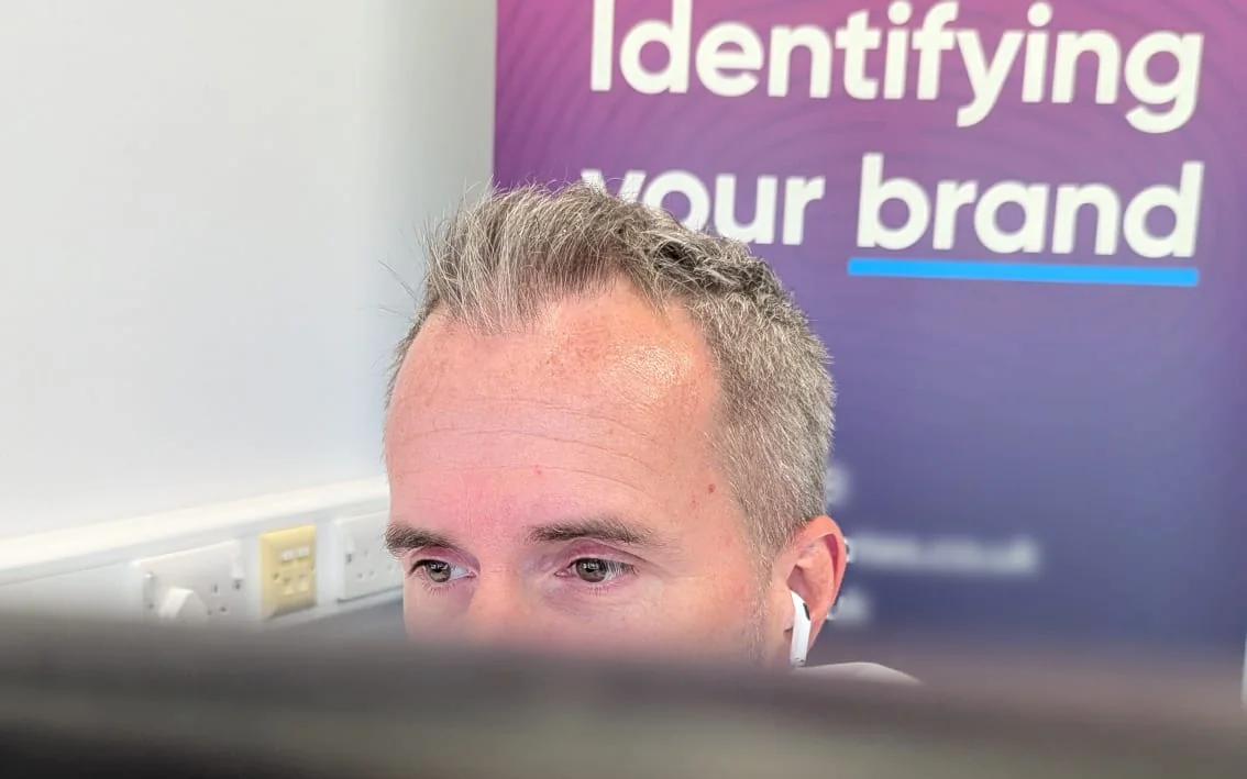 CJ Creates designer looking over his monitor in the Colchester studio, with the “Identifying your brand” banner behind