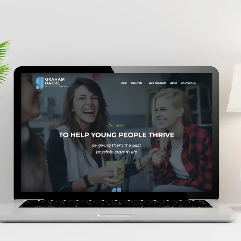Graham Dacre Charitable Foundation website home page on a laptop on a white desk beside a lamp and plant, with the headline 'To help young people thrive', designed by CJ Creates